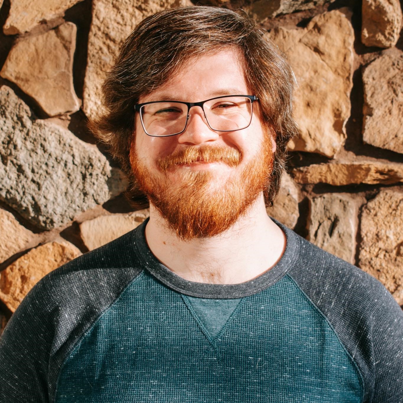 Headshot of a white man with shorter brown hair, glasses, and a red beard wearing a blue sweater in front of a stone background